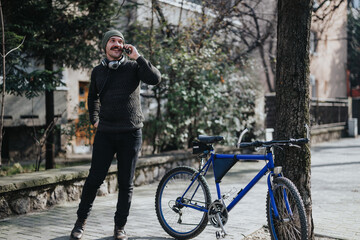 A cheerful young adult male enjoys a conversation on his smart phone while standing next to a blue bicycle on a sunny city street.