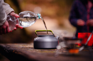 A person fills a camping kettle with clear water from a bottle, preparing for a warm drink in the outdoors, with unfocused figures in the background