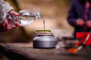 A person fills a camping kettle with clear water from a bottle, preparing for a warm drink in the...