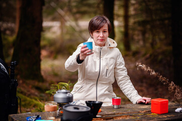 A woman warmly dressed in a cream jacket extends a mug, portraying a peaceful break with a camping...