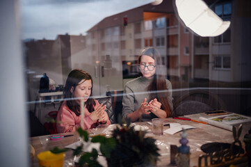 Through a window, a girl and a woman are seen focused on an art project together, cozy indoor creativity as dusk falls outside