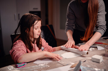 Intently focused, a mother assists her daughter with a clay craft project at home using rolling pin, showcasing a moment of shared concentration and educational interaction