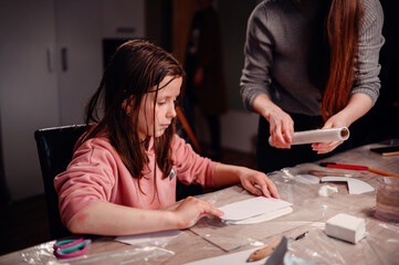 Fototapeta premium Intently focused, a mother assists her daughter with a clay craft project at home using rolling pin, showcasing a moment of shared concentration and educational interaction