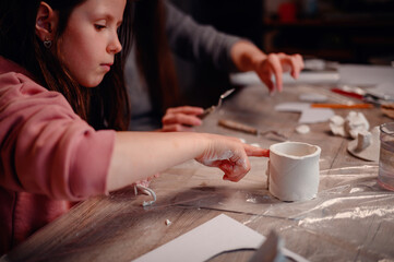 A child's hands carefully construct a clay house, with the focused guidance of a parent, symbolizing the foundational joy of building and learning together
