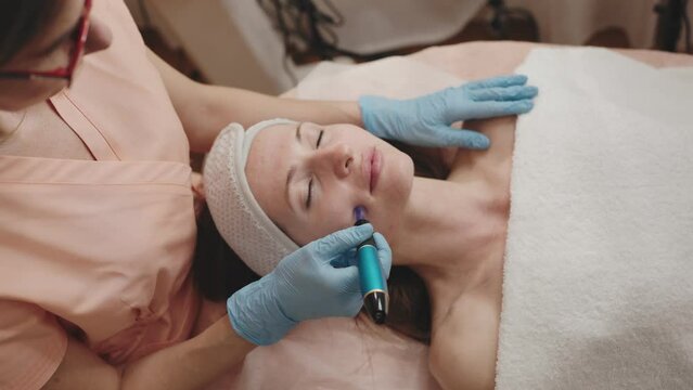 A skilled cosmetologist uses a derma pen for microneedling treatment on a relaxed female client in a beauty clinic.