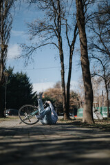 Obraz premium Photo capturing an unanticipated moment as a woman falls off her bike in a serene park setting, surrounded by bare trees and a clear sky.