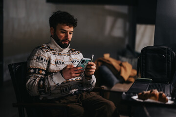 Focused man using smart phone in dimly lit room, with laptop and bag nearby, conveying a sense of business or travel.