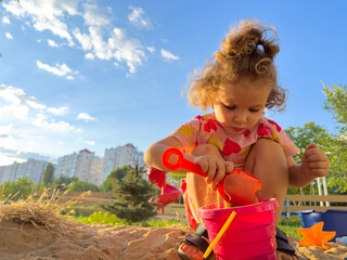 Little curly haired girl with toys playing in sandbox