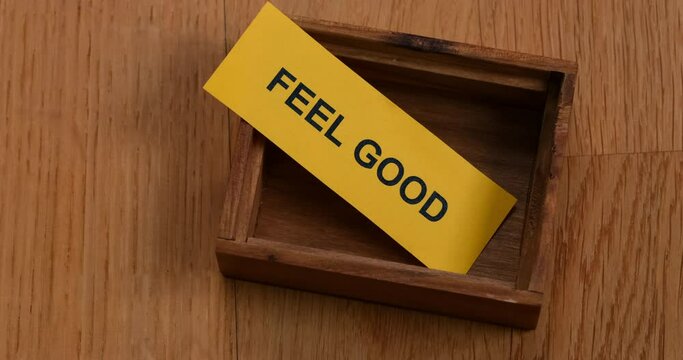 A yellow piece of paper with the phrase Feel Good on it lying inside of a small empty wooden box. Close up.