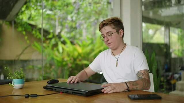 Tattooed transgender professional begins workday in eco-friendly modern office. Opens laptop, focuses on screen. Inclusive workplace promotes productivity, diversity. Slow motion.