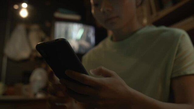 Medium close-up tilt shot of young Asian woman or man with short hair, in t-shirt sitting in dimly lit capsule flat and typing something on smartphone while resting at home after work