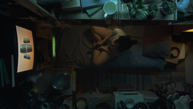Full top shot of Chinese or Korean girl sitting on bunk bed in cluttered micro flat, watching beauty commercial or reality show on TV, then lying down, while resting at home alone in evening