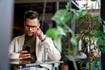 Handsome young businessman looking thoughtful while scrolling his mobile phone at coffee shop.