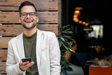 Smiling businessman with eyeglasses holding a smartphone while standing outside the Cafe.