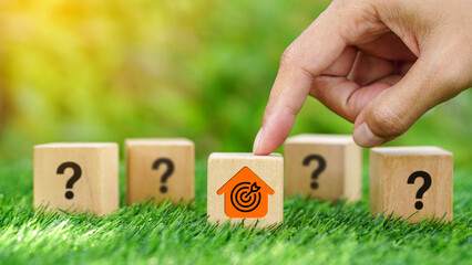 Hands point on house with target icon on wooden cube over green grass with blurred question mark...