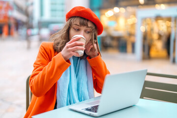 Fototapeta premium beautiful young woman in red coat and beret having coffee at the side cafe outside, using the laptop for studying, learning, working. lifestyle concept