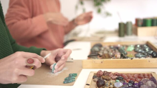 detail of a woman's hand sanding a gemstone, in the background a man weaving bracelets with the gems. Luminous photograph with green and salmon orange jumper.