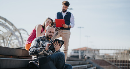 A trio of young professionals in casual business attire engaged in a collaborative discussion with a digital tablet, outdoors in a modern urban cityscape.