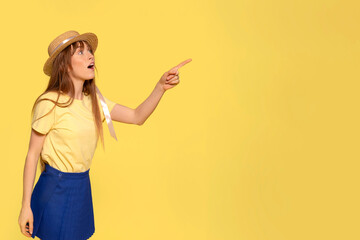 Surprised young redhead woman girl in a yellow t-shirt, blue skirt, and straw hat pointing her index fingers aside up on a yellow background with copy space.  Emotions lifestyle concept