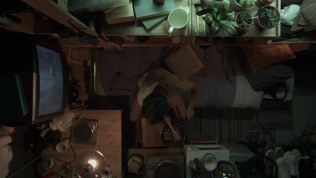 Full top shot of unrecognizable Asian male or female person sitting on bed in tiny apartment, putting stir-fry from pan into bowl, taking out chopsticks, preparing to eat dinner and watch movie on TV