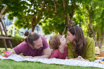 Parents leaning in to kiss young boy on picnic blanket, amidst a spread of snacks, portraying family affection and shared joy. Mother and father bestow peck on cheek of boy amidst outdoor feast - obrazy, fototapety, plakaty