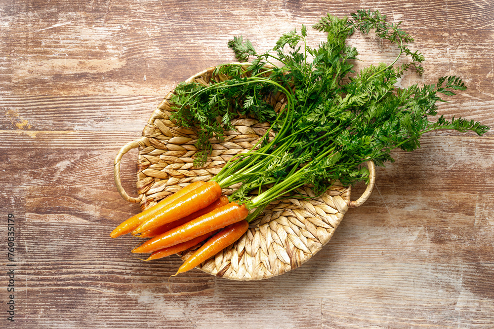 Sticker fresh organic farmer carrots bunch with green tops in a basket on wooden rustic kitchen table, top d