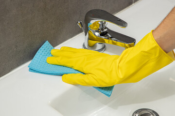 Close up of a hand in a yellow protective glove cleaning or wiping a modern, white, ceramic sink or washbasin, with a blue cloth and a shiny, chrome tap or faucet.