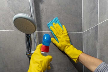Close up of hands wearing yellow gloves cleaning a shower cubicle with a blue cloth and a bottle of...