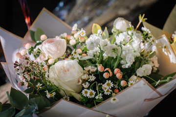 Valmiera, Latvia - March 7, 2024 - a bouquet of white roses, chamomile, and other flowers with green leaves wrapped in brown paper with a windowpane pattern.