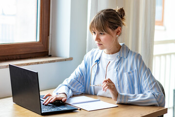 Young woman freelancer in casual clothes sitting at table using laptop working online at home