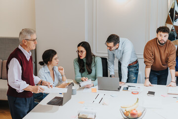 A multi-generational group of professionals discuss strategy around a table with laptops,...