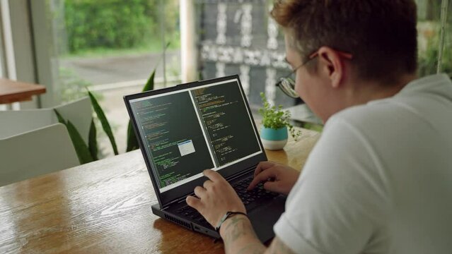 Transgender programmer codes on laptop in coworking space, focused on debugging software. Gender diverse specialist works in tech office, writes innovative applications in vibrant startup hub. Slowmo