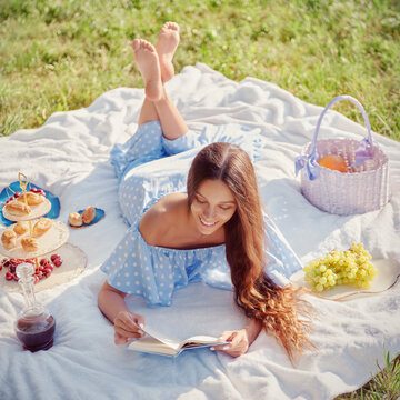 Picnic scene: sweet smiling girl lying on plaid and reading book in garden. Outdoor portrait of beautiful happy young woman in blue dress