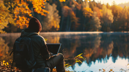 Man with a backpack and a laptop sits on the shore of a mountain lake in autumn - obrazy, fototapety, plakaty