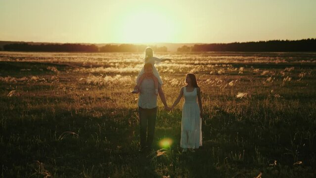 Father Carries Daughter On Shoulders Holding Hand Of Wife Walking In Field. Family Gets Home Together To Celebrate Mum Birthday. Family Discusses Plans For Future Telling Funny Jokes At Sunset