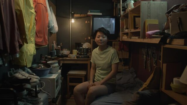 Full portrait shot of young Chinese woman sitting on bunk bed in cluttered shabby micro rental apartment and looking at camera, TV playing movie in background, microwave, possessions in bags on wall