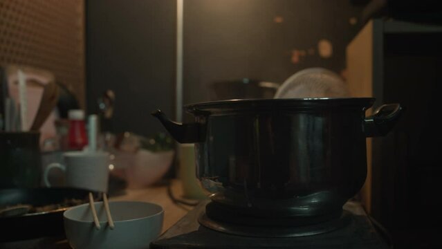 Medium no people shot of steaming pot on small electric stove, bowl with chopsticks, frying pan with food and crockery, on table in shabby dimly lit capsule flat in China or Hong KongMedium no people 