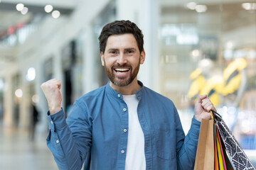 Cheerful bearded man in a mall holding colorful shopping bags, feeling victorious and happy about...