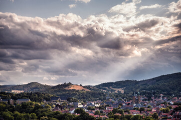 Obraz premium A dynamic cloudscape crowns a peaceful town, nestled among lush hills, with rays of sunlight breaking through to highlight its charm