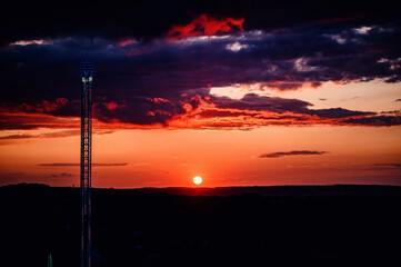 The sun sets in fiery splendor behind a towering ride, its silhouette etching a striking contrast...