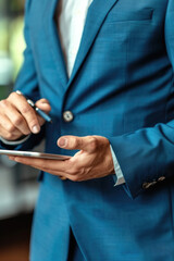 A man dressed in a blue suit holding a tablet device. He appears focused on the screen, standing confidently