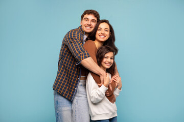 Family portrait. Cheerful father, mother and preteen girl posing isolated on blue studio background, loving parents hugging child girl and smiling