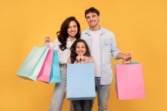 European family of three shopping together, posing with colorful shopper bags and smiling over yellow studio background. Seasonal sales concept