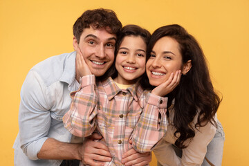 Closeup portrait of european family of three with preteen daughter, parents and their child girl embracing and posing smiling at camera on yellow background