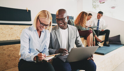 Two diverse business people laughing together while sitting and working on some notes. Meanwhile, their three colleagues sit in the background and concentrate on working together.