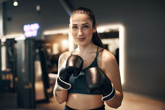 Fit young woman wearing a tank top and boxing gloves smiling while standing in a ready stance during a self defence and workout session in a gym