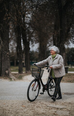 Mature female retiree pushing a bicycle on a peaceful path in a scenic park, embracing an active lifestyle.