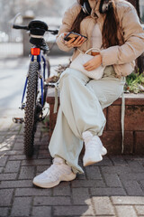 A trendy young adult rests on a city bench with her bicycle, using a smart phone and enjoying music...