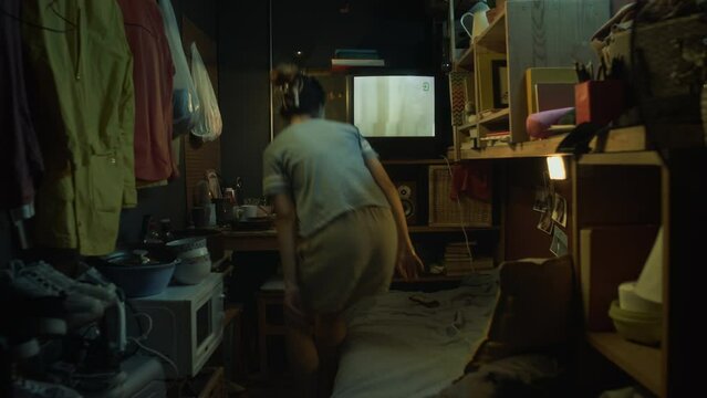 Full shot of Chinese girl in casual t-shirt and shorts walking into tiny capsule apartment, turning on TV, laying down on bunk bed to watch, while relaxing at home after work