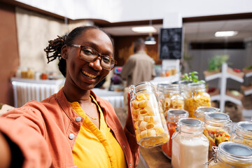 In bio-food shop, black woman wearing spectacles holds glass container loaded with spaghetti....